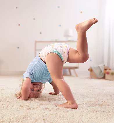 Niño pequeño feliz en pañal-braguita Dodot haciendo una vertical sobre una alfombra suave, sonriendo a la cámara.