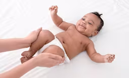 A baby lying on their back wearing a diaper. A caregiver has their hands on the baby’s legs