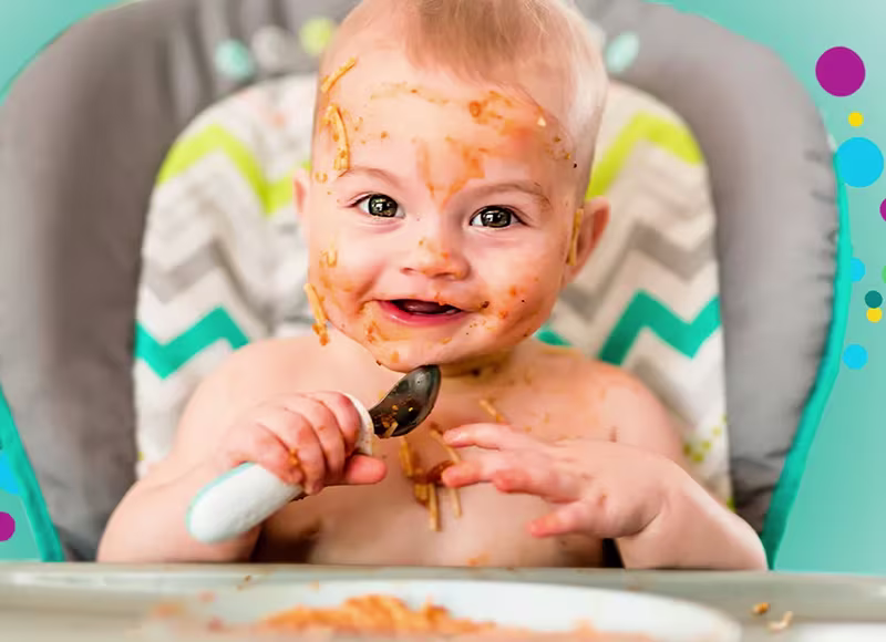 Χαμογελαστό μωρό σε ψηλό καρεκλάκι, με το πρόσωπο καλυμμένο με σάλτσα σπαγγέτι, κρατώντας ένα κουτάλι Χαμογελαστό μωρό σε ψηλό καρεκλάκι, με το πρόσωπο καλυμμένο με σάλτσα σπαγγέτι, κρατώντας ένα κουτάλι