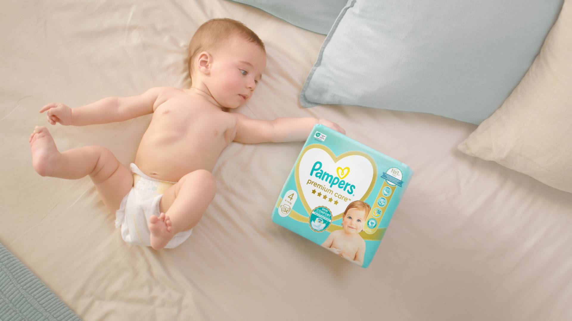 Baba a hátán fekszik, Pampers Premium Care pelenkát visel, és ujjhegyével megérinti a Pampers Premium Care csomagolást