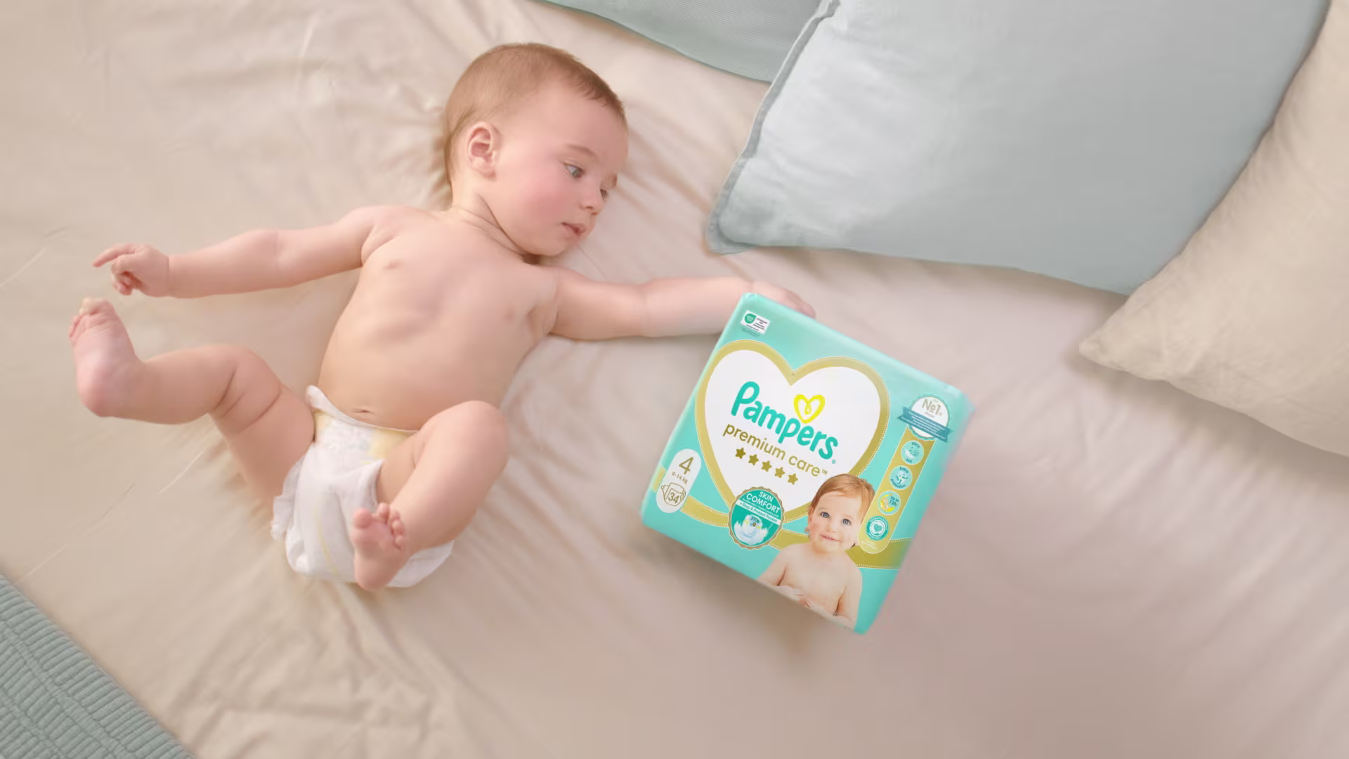 Baba a hátán fekszik, Pampers Premium Care pelenkát visel, és ujjhegyével megérinti a Pampers Premium Care csomagolást