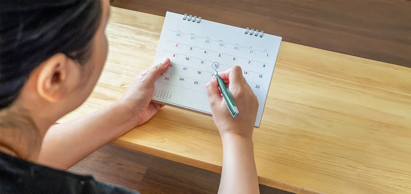 Personne assise à une table, marquant une date sur un calendrier posé devant elle.