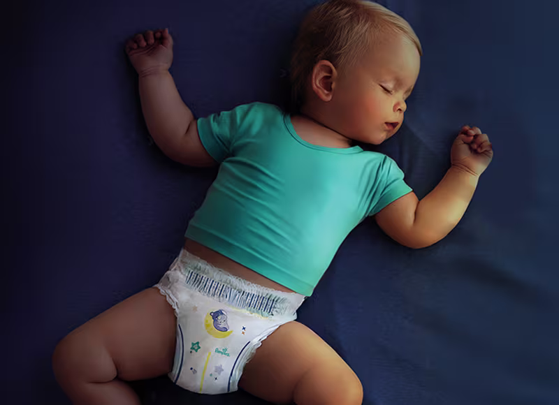 Dziecko ubrane w turkusową koszulkę i pieluszkę Pampers spokojnie śpi na ciemnoniebieskiej powierzchni.
