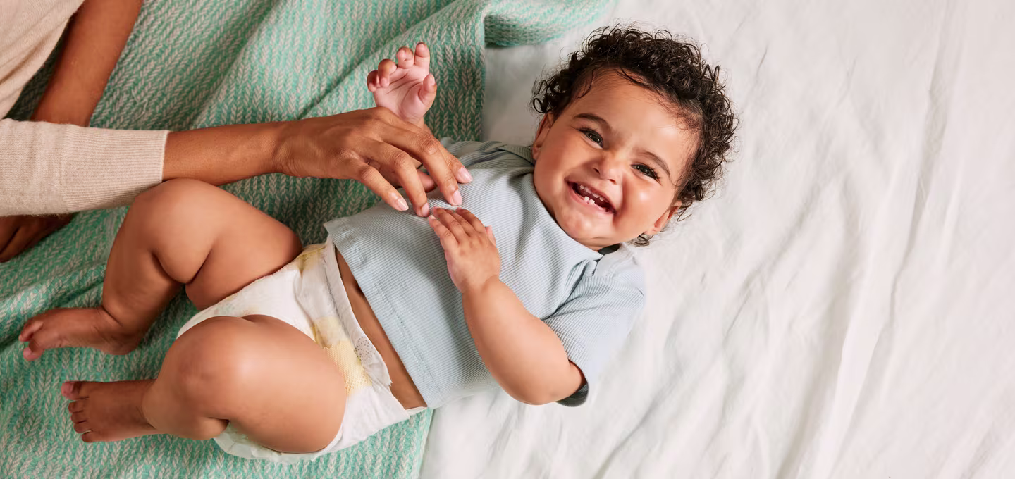 Strahlend lächelndes Baby in einer Windel liegt auf dem Bett, während ein Erwachsener liebevoll mit ihm spielt.