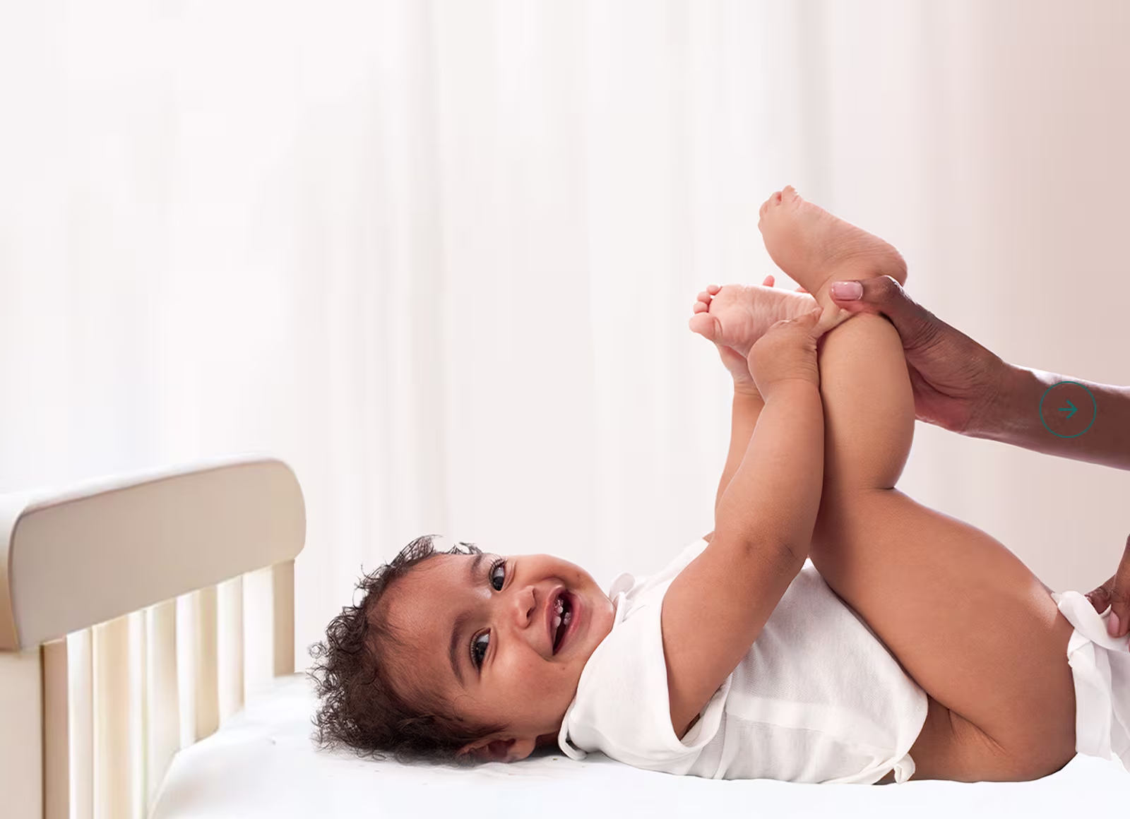 A baby lies on their back on a white surface, holding their feet up while a parent is using a wipe to clean the baby’s bottom.