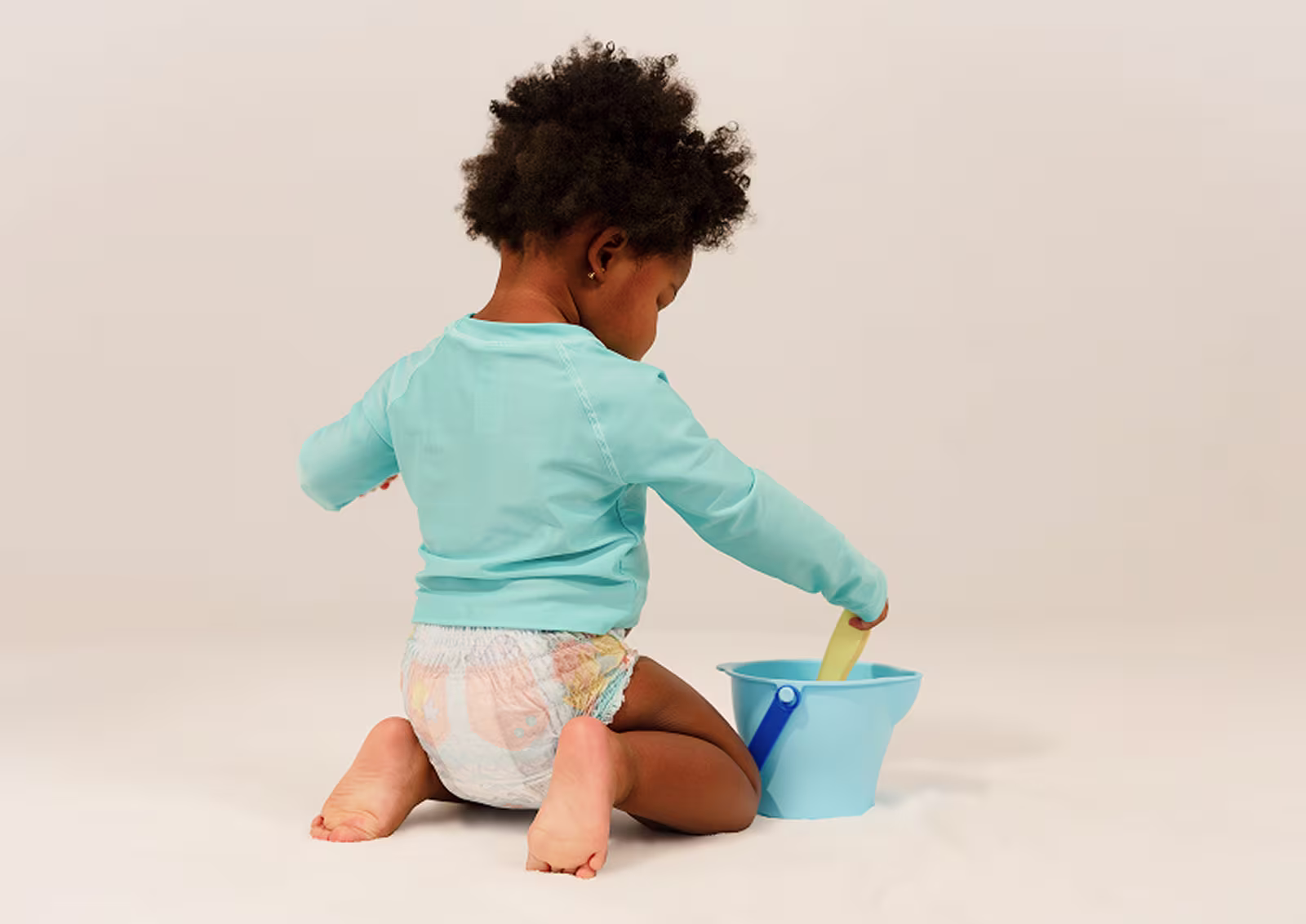 Ein Kleinkind, das Pampers Splashers und ein türkisfarbenes Schwimmshirt trägt, kniet beim Spielen mit einer Schaufel und einem Eimer.