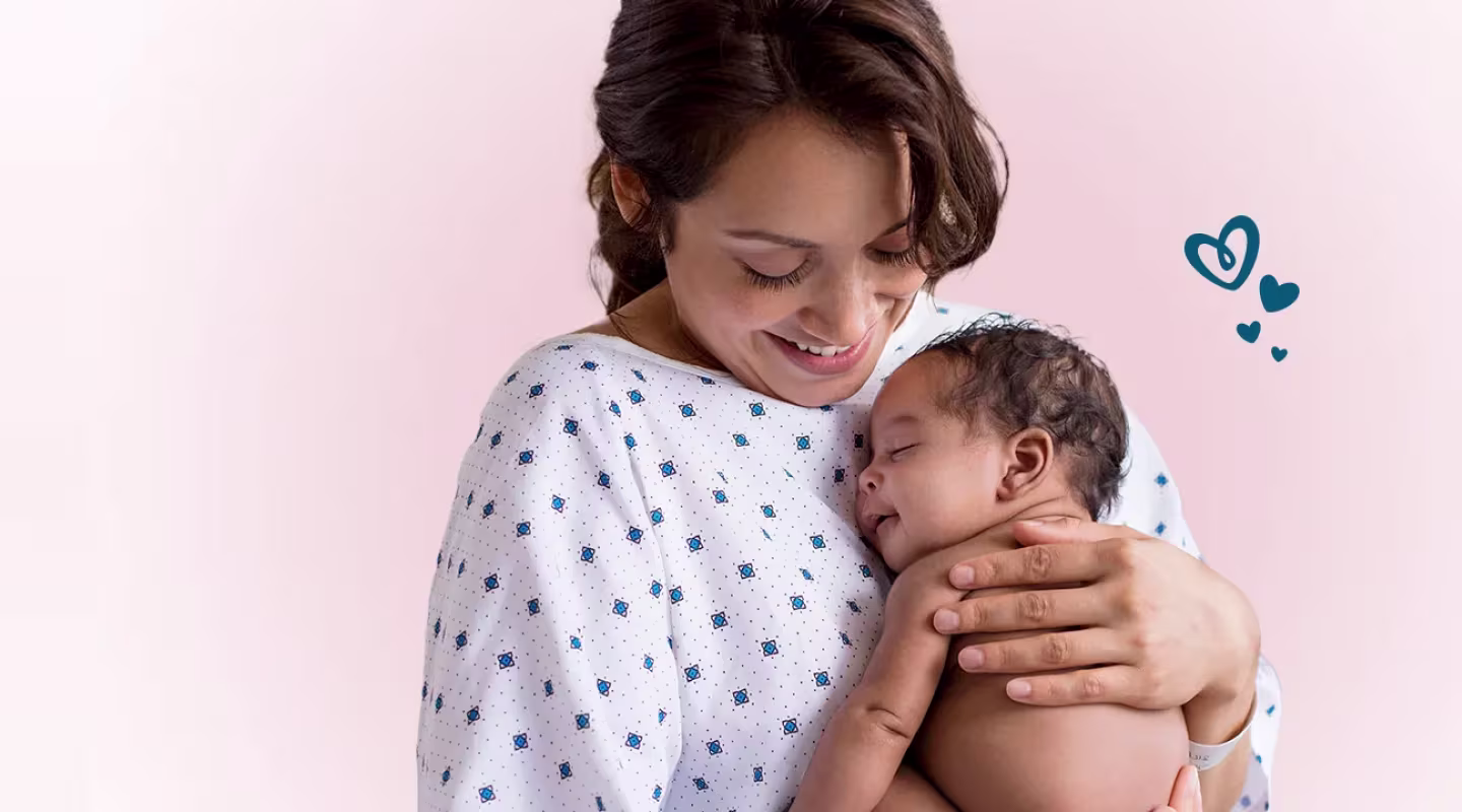 Una madre sonriente vestida con una bata de hospital sostiene a un bebé recién nacido dormido cerca de su pecho.