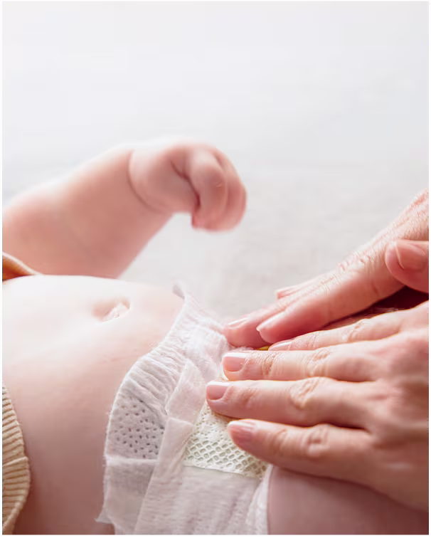 A closeup of a baby lying down during a diaper change, wearing a light yellow shirt. The hands of a parent are shown gently pressing the diaper tabs down.