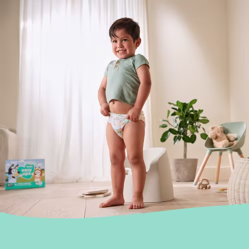 Toddler smiling while pulling up training pants beside a potty chair, representing a child-led approach to potty training