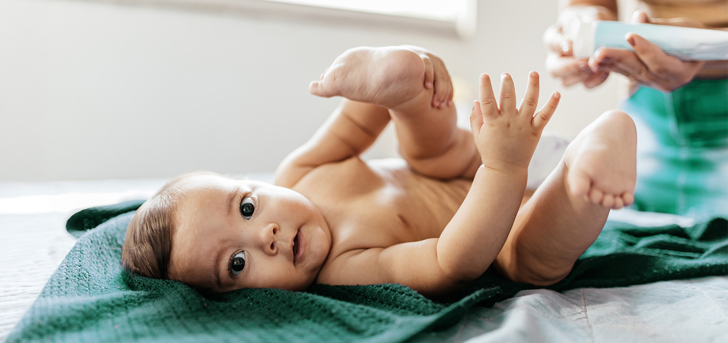 Baby liegt auf einer Decke und hebt spielerisch Beine und Arme, während ein Erwachsener im Hintergrund eine frische Windel bereithält.
