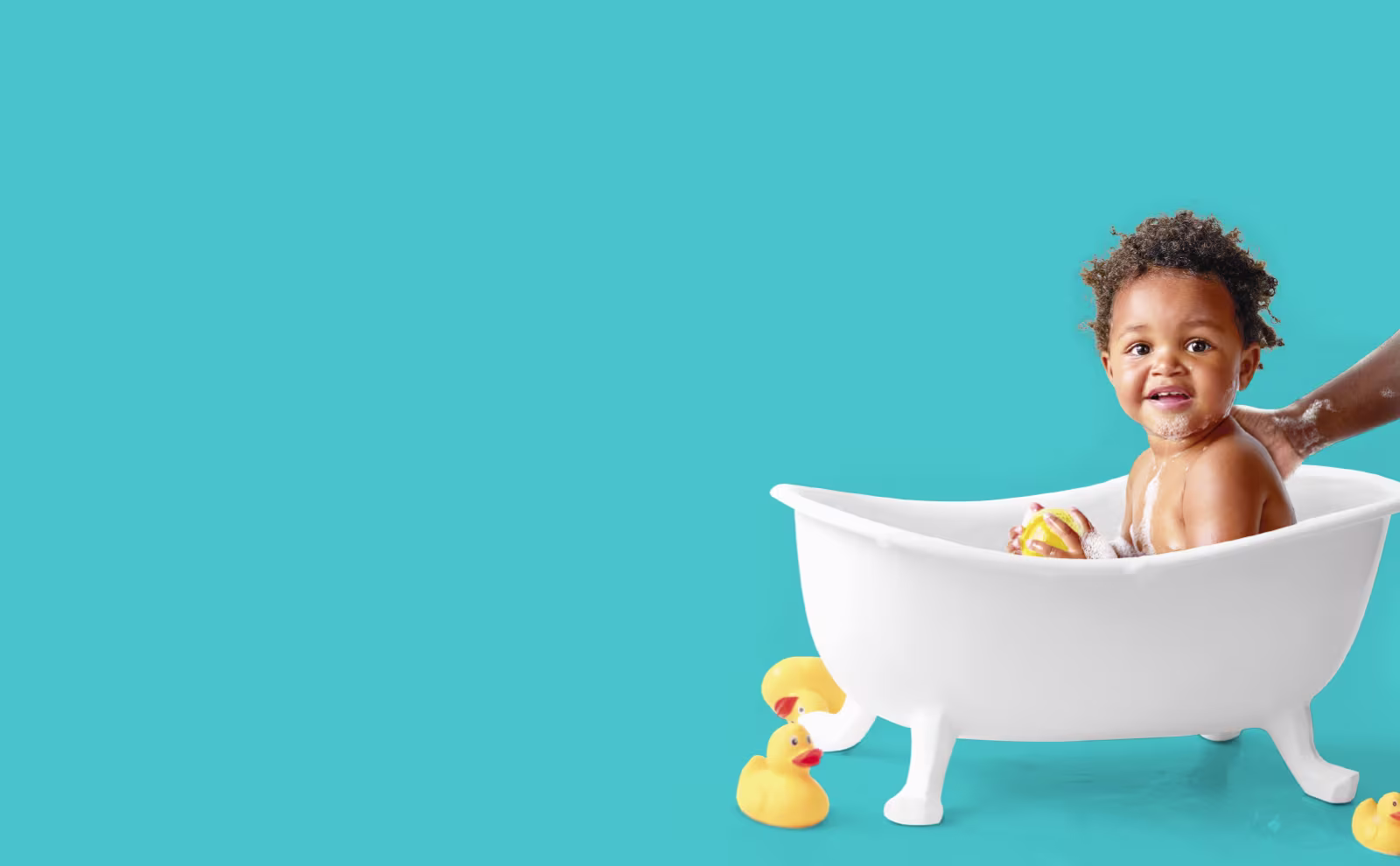 Un copil mic și fericit stă într-o cădiță albă. Mâna unui adult îi spală ușor spatele, în timp ce niște rațe de cauciuc galbene stau pe podea lângă el.