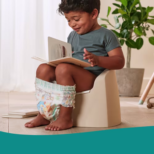 Toddler sitting on a potty chair reading a book with training pants around ankles, illustrating the 3-day potty training method.