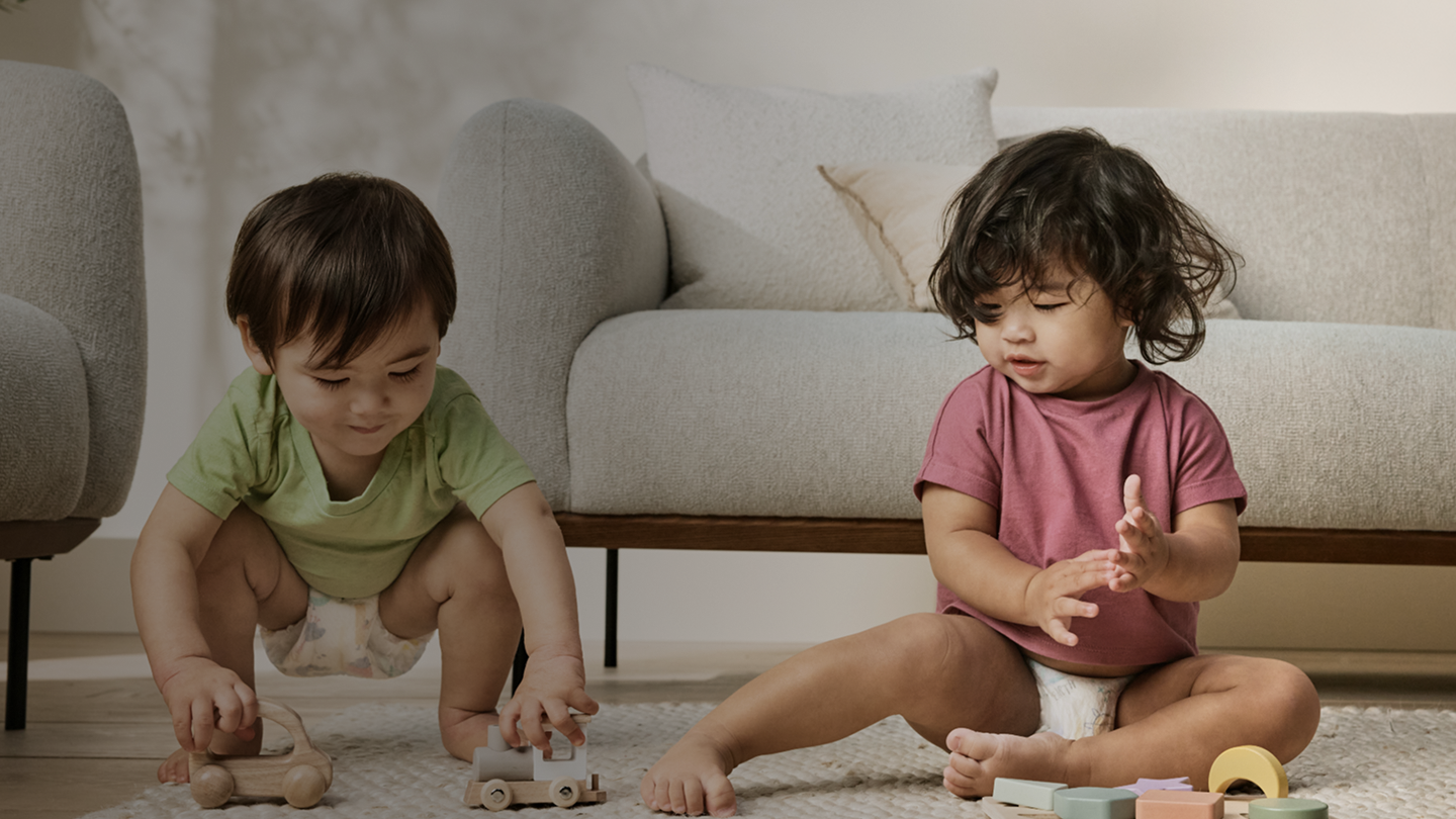 Two babies playing with wood toys on floor carpet. A sofa is behind them 