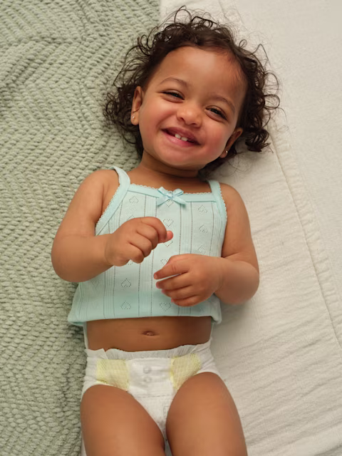 Bebé acostado sobre mantas de colores neutros usando pañales Pampers Swaddlers y una camiseta sin mangas color verde azulado.