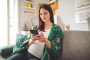 Pregnant woman in a green floral robe sits on a couch, smiling at her phone in a cozy living room.