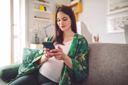 Pregnant woman in a green floral robe sits on a couch, smiling at her phone in a cozy living room.