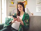 Pregnant woman in a green floral robe sits on a couch, smiling at her phone in a cozy living room.