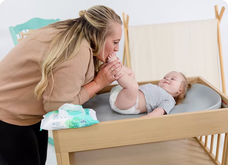 A loving mother gently kisses her baby's tiny feet during a diaper change, capturing a tender moment of care and affection. Play pen in the background and Aqua Pure wipes pack resting on the edge of the changing table