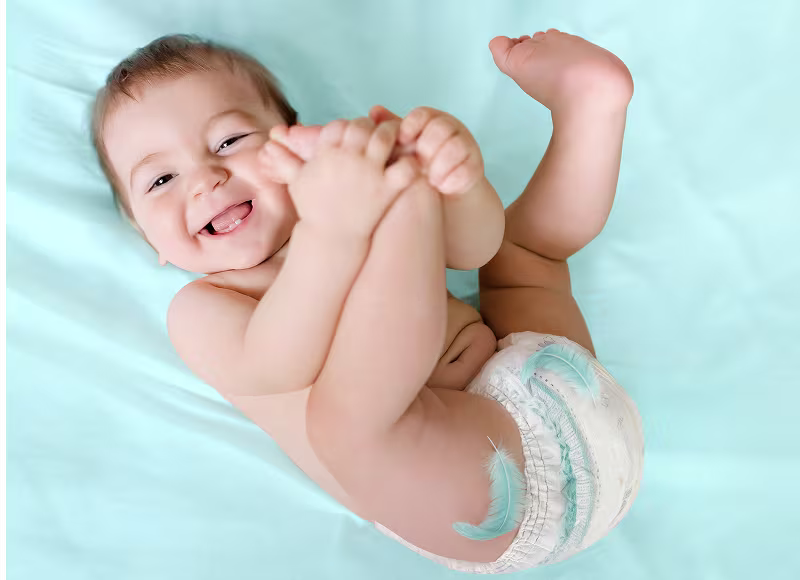 En glad baby som ligger på ryggen på et lyseblått laken, holder i føttene sine og har på seg en Pampers bleie.