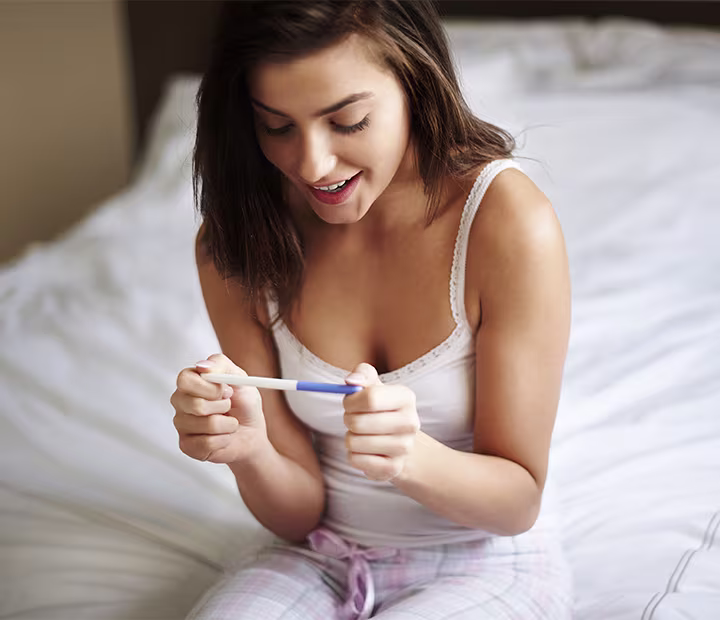 Woman on a bed joyfully looking at a pregnancy test.