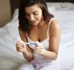 Woman on a bed joyfully looking at a pregnancy test.