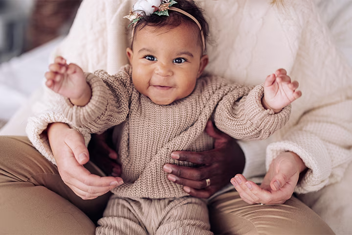 O fetiță fericită stă în brațele mamei sale, care o cuprinde  cu mâinile.​
