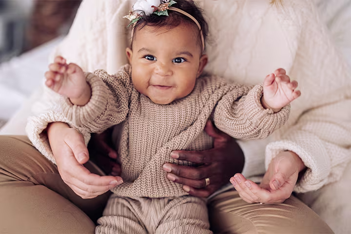 Pampers Article Images 720x480 girls names 01 O fetiță fericită stă în brațele mamei sale, care o cuprinde cu mâinile.