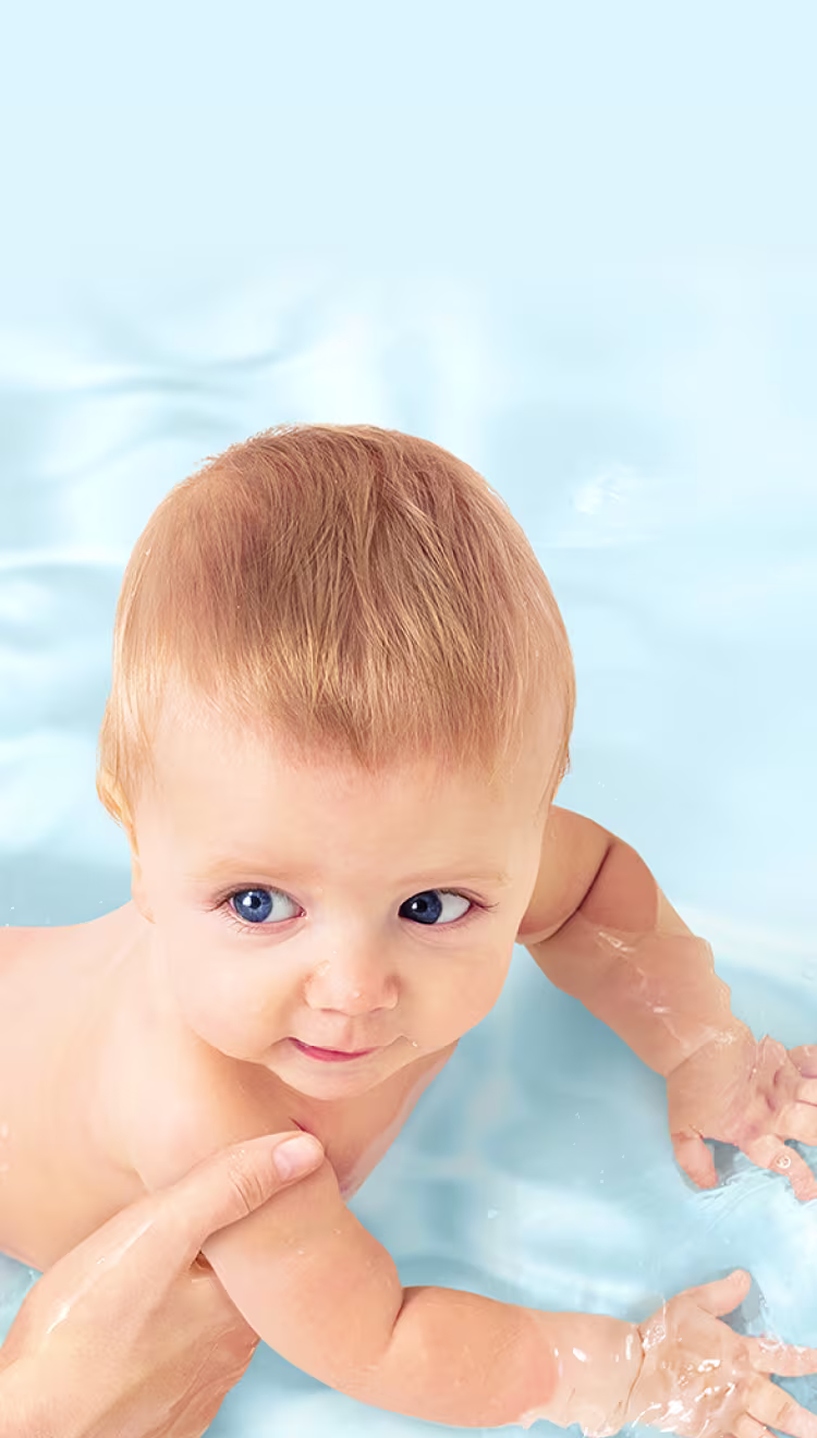 Una vista aérea de un bebé siendo sostenido suavemente en agua clara y azul por la mano de un adulto.