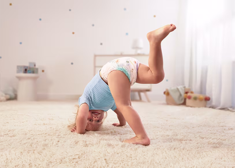 Glad liten i Pampers Baby-Dry blöjbyxor som gör en handstående på en fluffig matta och ler mot kameran.