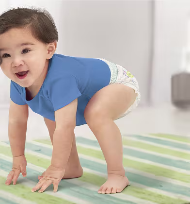 Um bebé com uma fralda-cueca e uma camisola azul, apoiado nas mãos e joelhos sobre um tapete às riscas, a sorrir para o espectador.