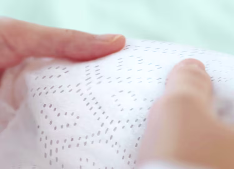 Eine Nahaufnahme zeigt zwei Hände, die sanft das Pampers Windelinnenvlies halten, wobei die Struktur und die Weichheit verdeutlicht werden.