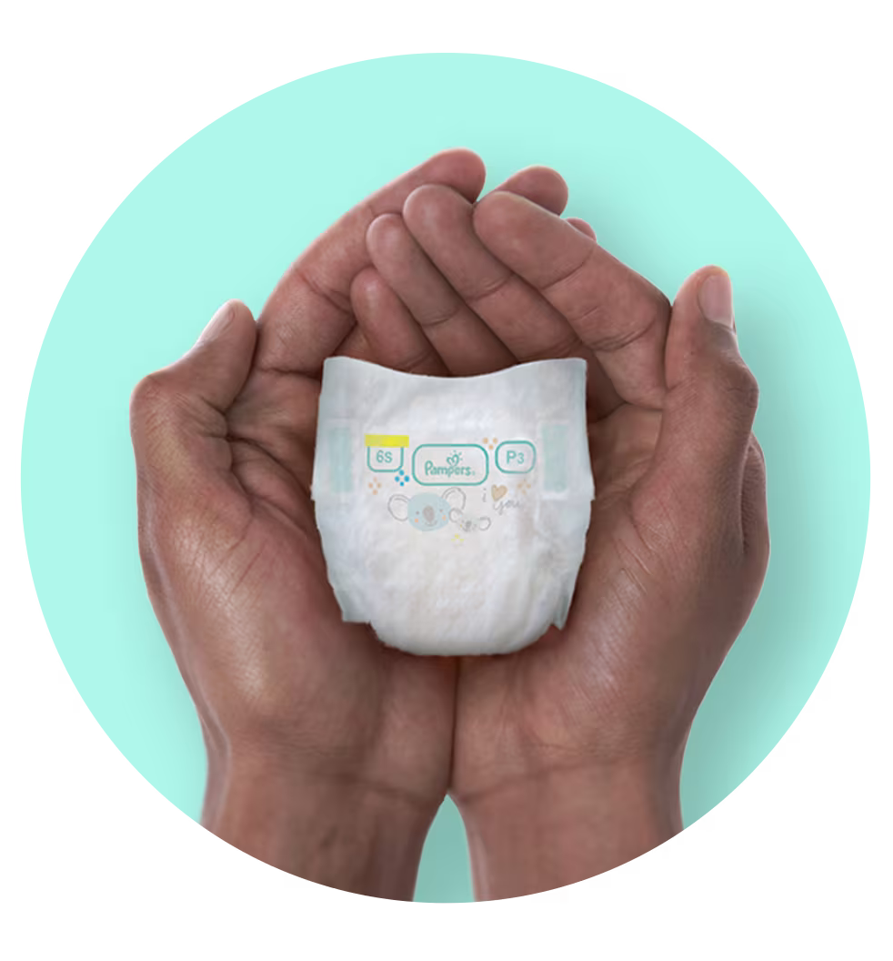 Adult hands gently cupping a tiny Pampers nappy against a soft blue background, emphasising the miniature size designed for premature babies.