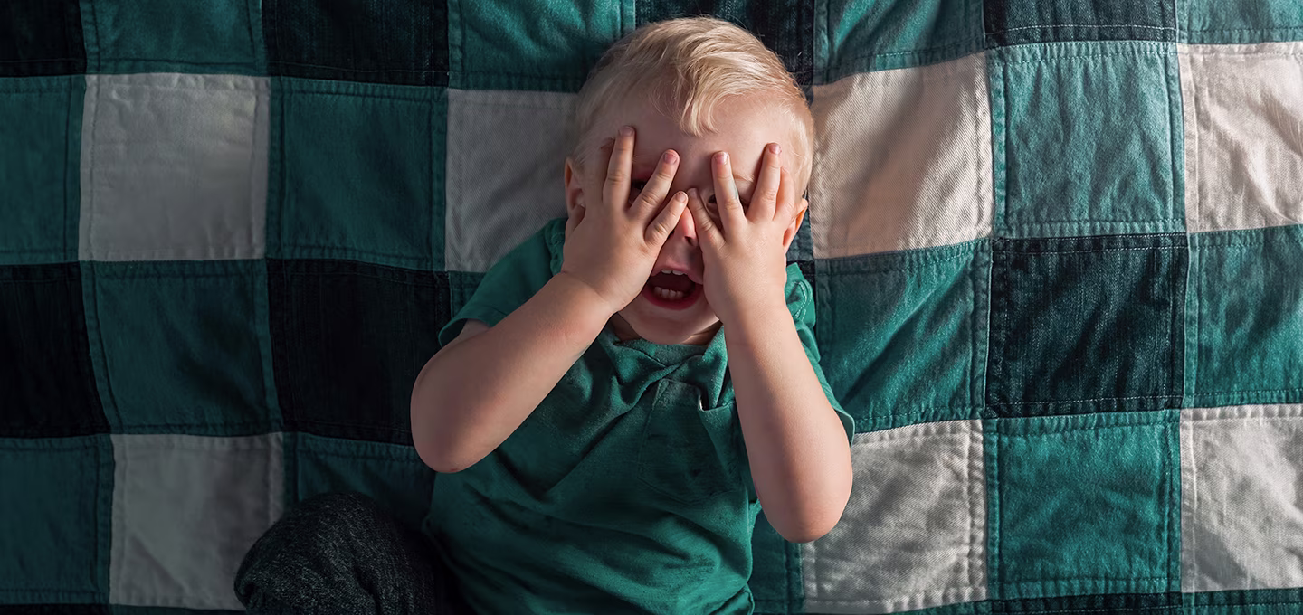 Toddler on a bed covering their face with both hands, appearing upset or distressed during a moment of sleep regression.