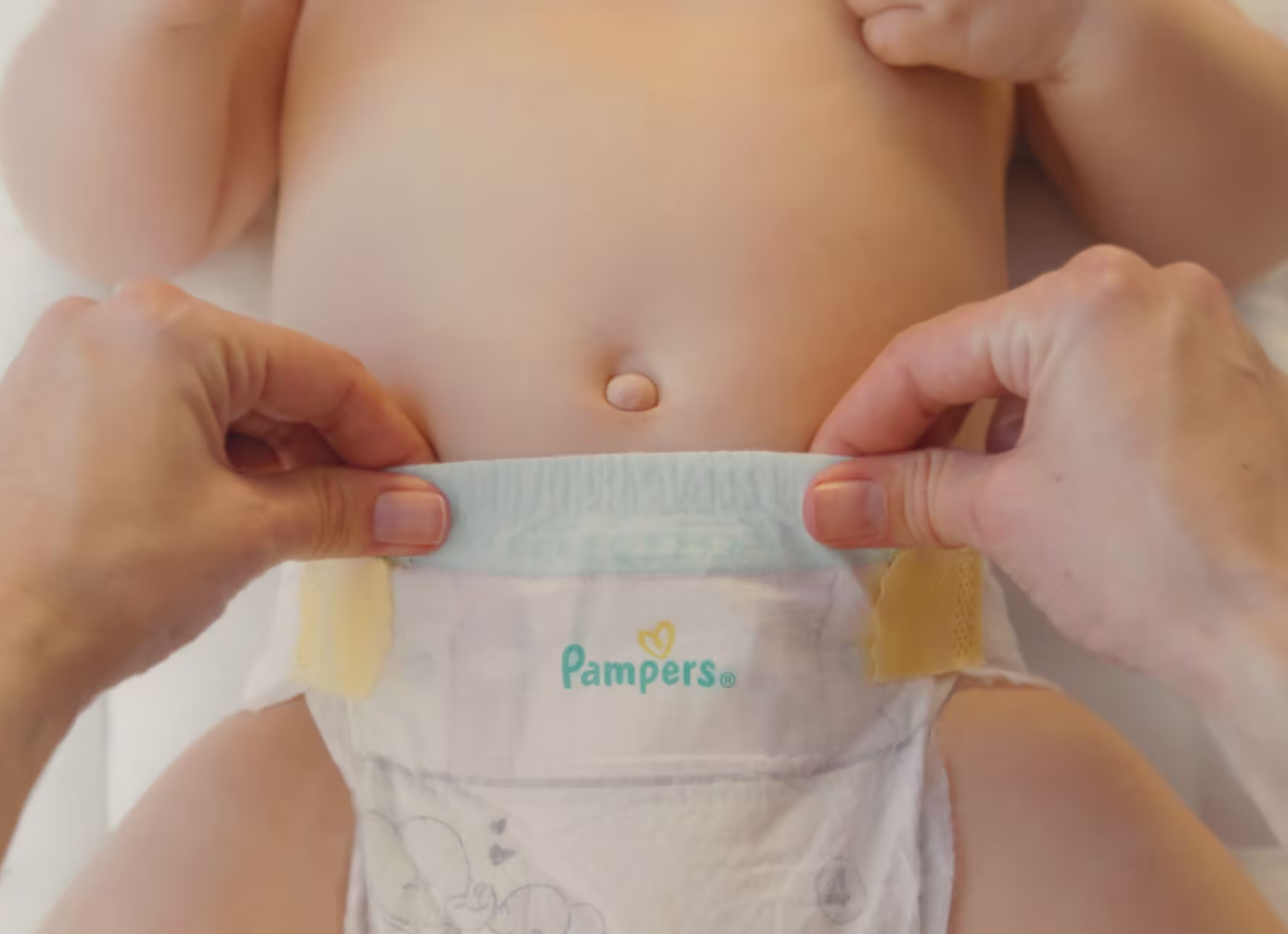 Een close-up toont twee handen die voorzichtig aan de buikband van een Pampers-luier trekken om de soepelheid en bescherming van de buikband te demonstreren.