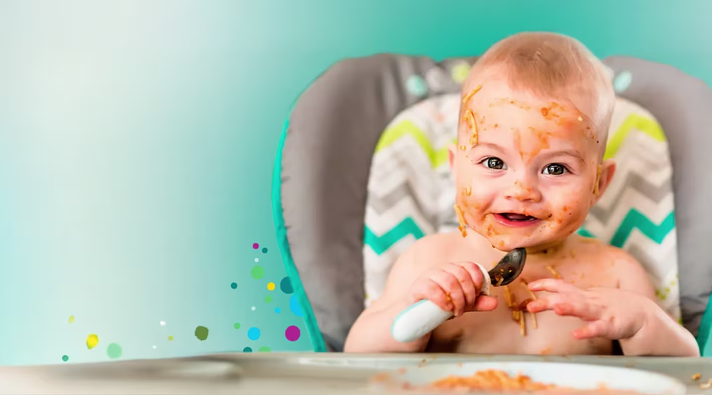 Χαμογελαστό μωρό σε καρεκλάκι φαγητού, με το πρόσωπο καλυμμένο με σάλτσα σπαγγέτι, κρατώντας ένα κουτάλι