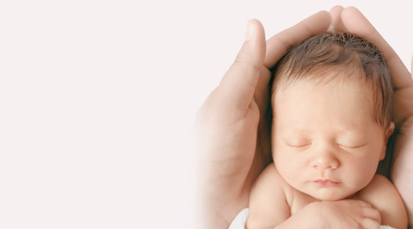 A pair of gentle hands carefully cradles the head of a sleeping newborn baby against a light background.