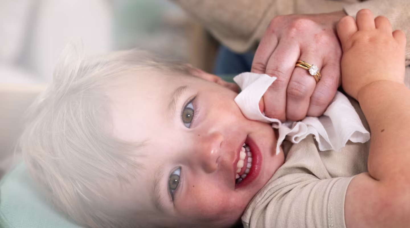 Un niño pequeño con cabello castaño y ojos marrones claros mira hacia arriba desde un cambiador verde, sonriendo mientras las manos de una mujer le limpian suavemente la cara con una toallita blanca.
