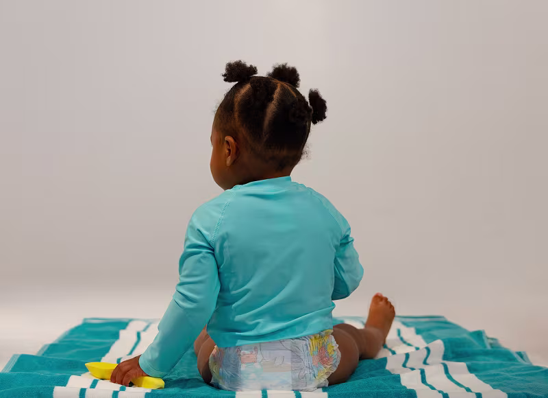 Uma criança pequena, vestida com calções Dodot Splashers e um fato de banho turquesa, está sentada de costas para a câmara.