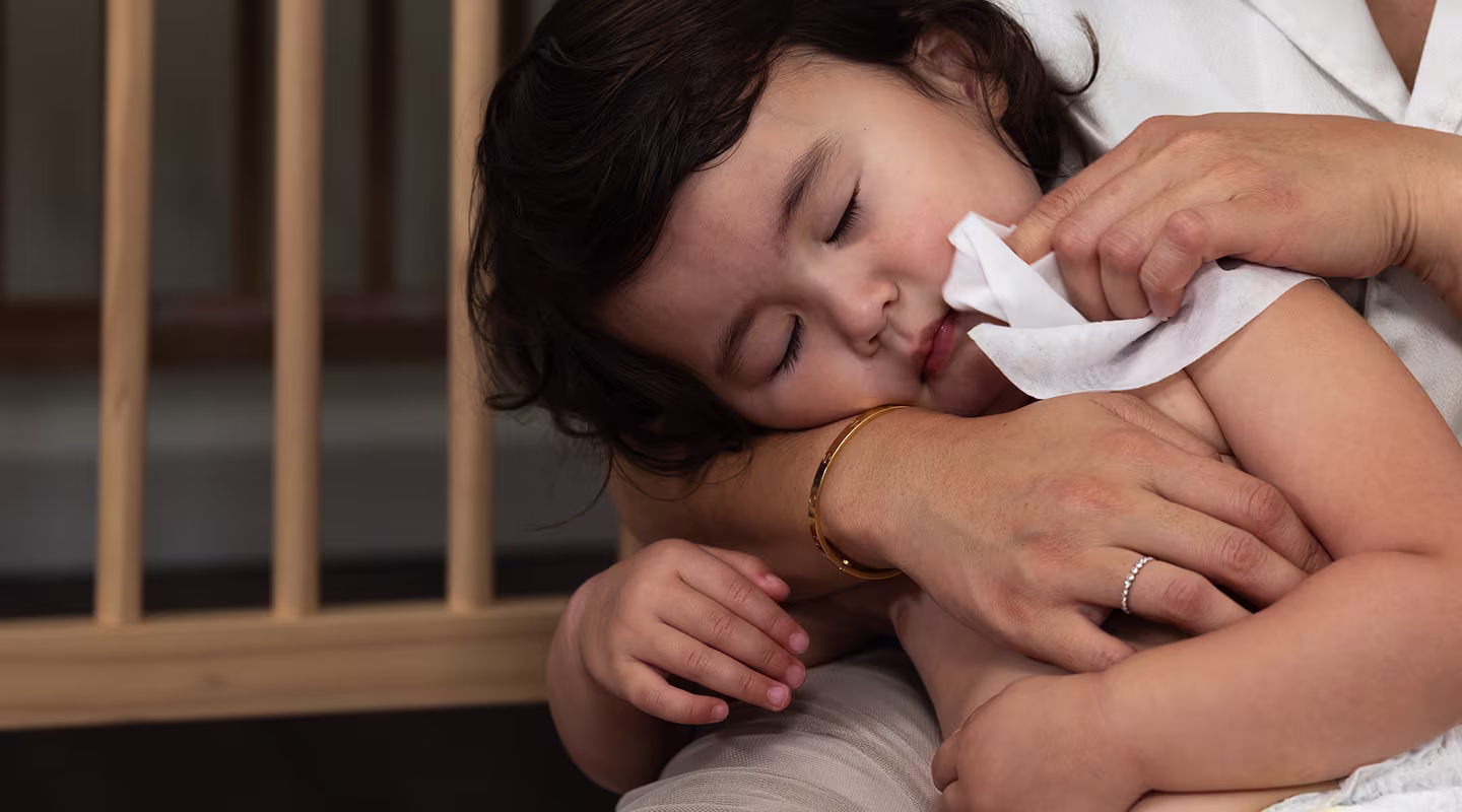 A gentle mother embraces her child while sitting beside a crib. She lovingly wipes her sleeping daughter's face with a soft baby wipe, ensuring her skin stays clean and fresh.