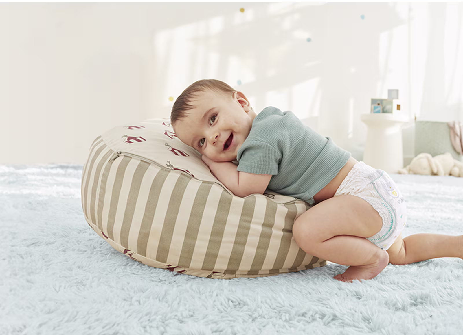 Een baby die een Pampers® Premium Protection™ Pants draagt, kijkt vrolijk naar de camera en leunt op een gestreept kussen.