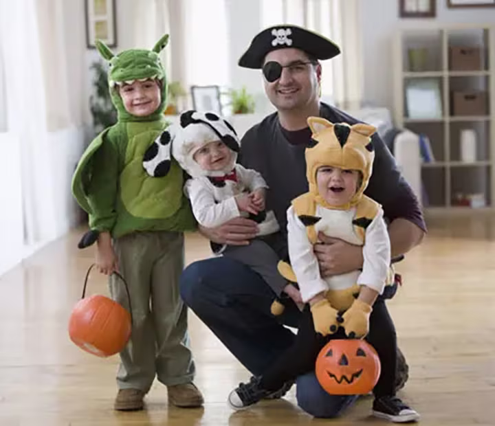 Family in cute animal and pirate costumes with pumpkin buckets, smiling indoors.