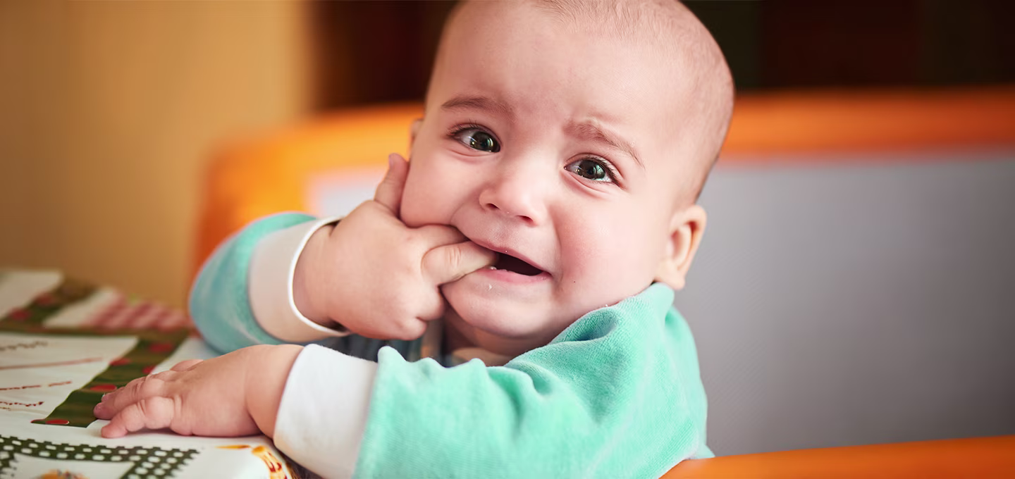 Baby with watery eyes and a worried expression chewing on fingers, showing common signs of discomfort that may occur during teething.
