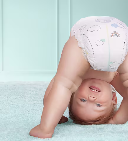 Ein fröhliches Baby in einer Windel steht lächelnd kopfüber auf einem weichen, hellblauen Teppich. Die Windel ist mit niedlichen Wolken- und Regenbogenmotiven verziert. Im Hintergrund ist eine Wand in sanftem Blaugrün zu sehen.
