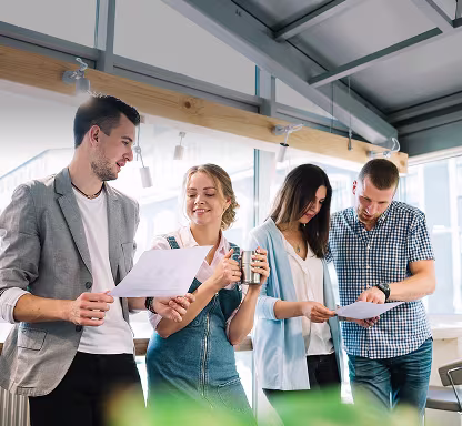 Four coworkers stand in a bright office, smiling and discussing papers and notes together.