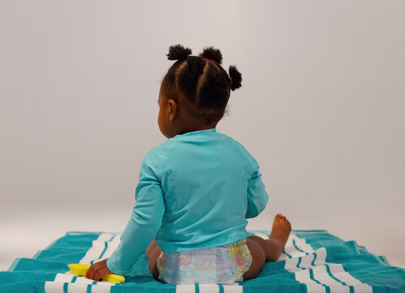 Маленька дитина в підгузках Pampers Splashers стоїть на колінах, граючись лопаткою та відерцем. Маленька дитина в підгузках Pampers Splashers стоїть на колінах, граючись лопаткою та відерцем.