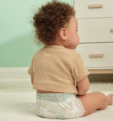 A close up of the back of a toddler sitting in their nursery while wearing a Pampers Swaddlers 360 pull on diaper and neutral color tshirt. A white dresser stands in the background
