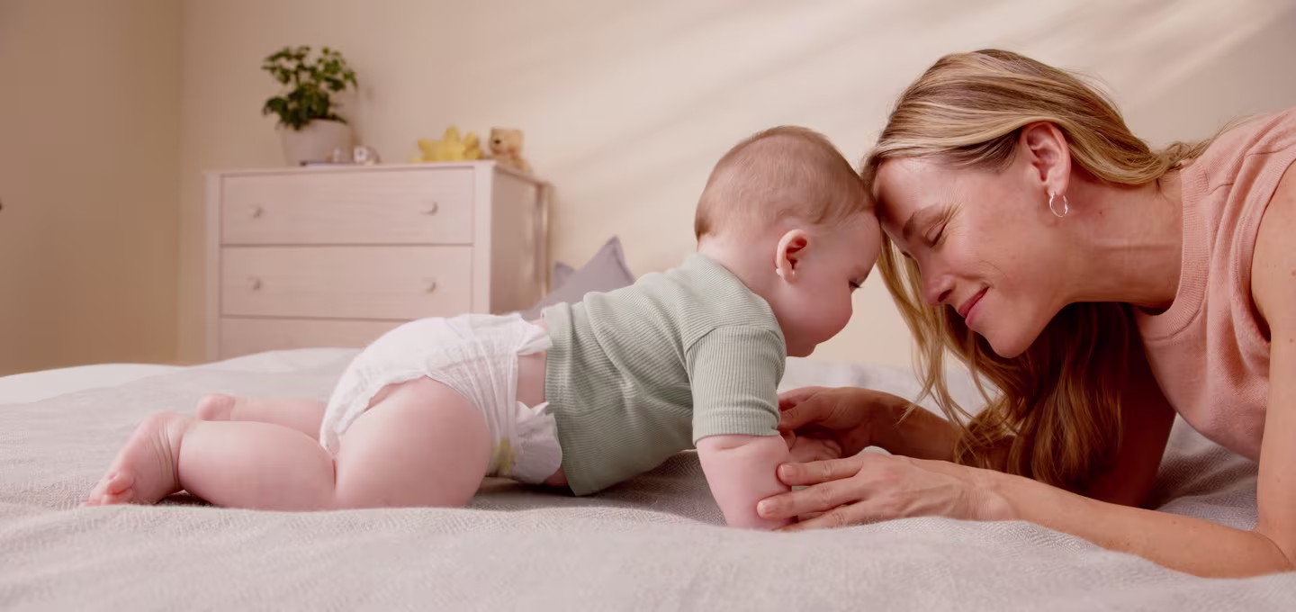 Mother and baby touch foreheads while playing on a bed in a bright bedroom.