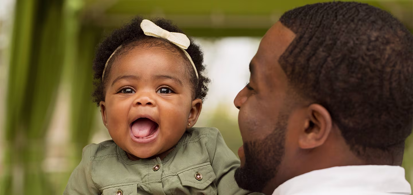 Smiling baby with a bow headband laughs while being held by a parent outdoors.