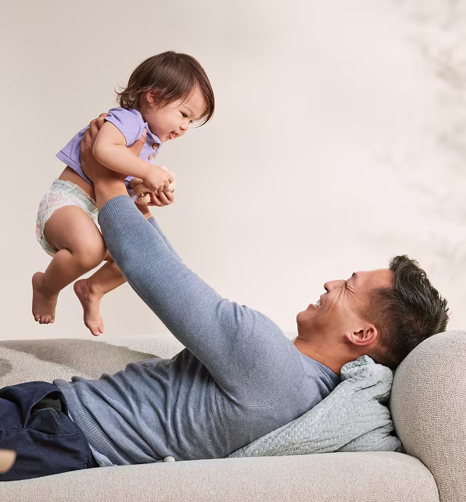 Father wearing a grey long-sleeve shirt smiling at baby that he is holding in his arms in the air. Baby wearing Pampers diapers smiling