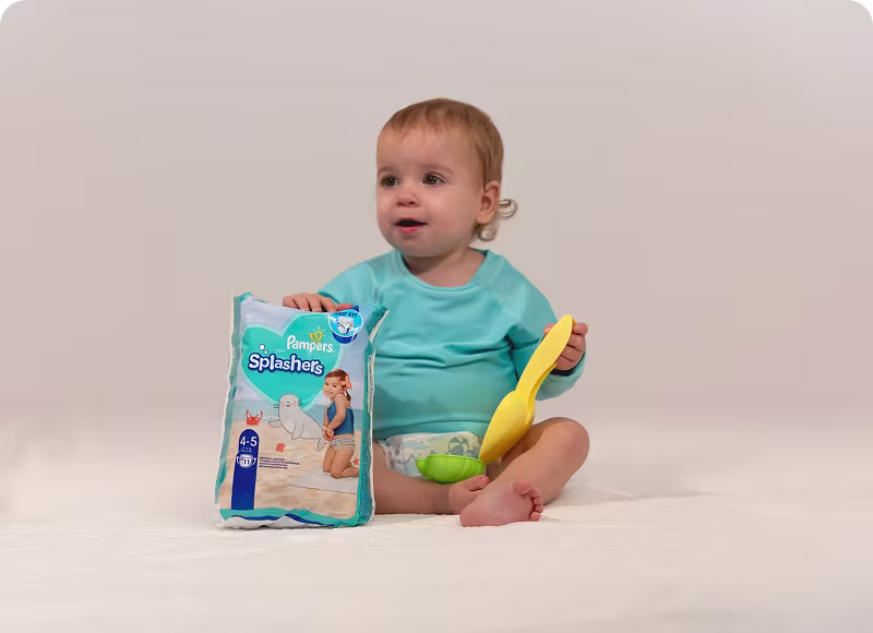 Un bebé alegre se sienta orgulloso sosteniendo un paquete de Pampers Splashers y una pala para arena, ¡listo para un día divertido en la alberca!